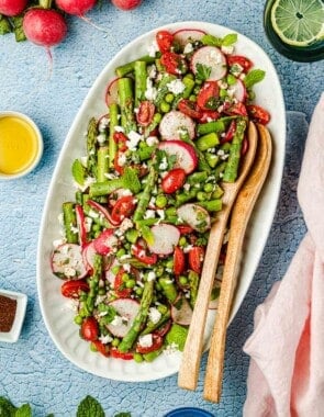 Asparagus salad on a large white serving platter with two wooden serving spoons and lemon water on the side.