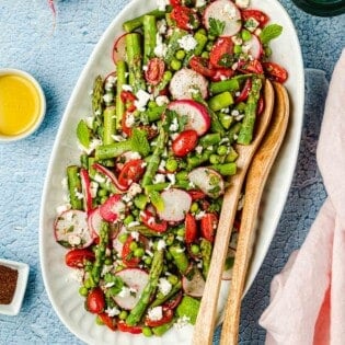Asparagus salad on a large white serving platter with two wooden serving spoons and lemon water on the side.