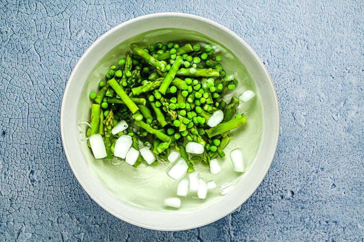 Blanched peas and asparagus in a bowl with ice water.