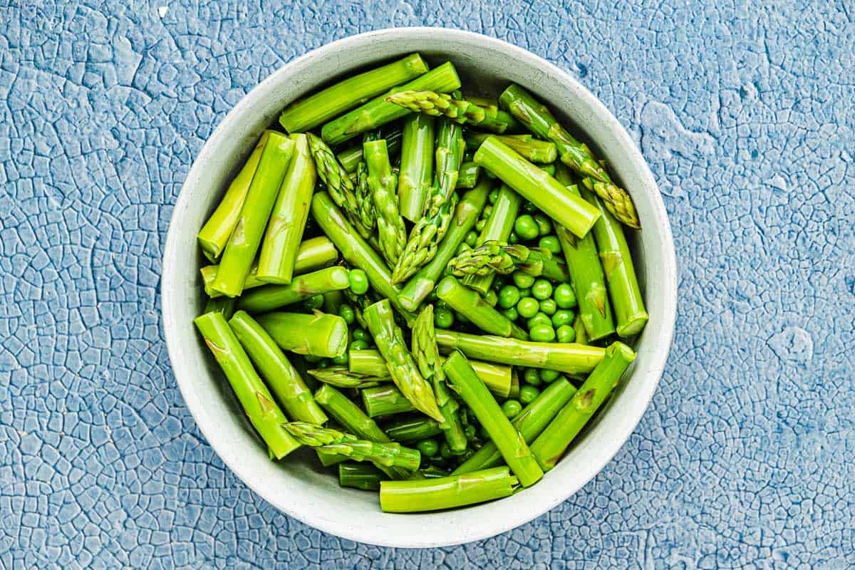 Sliced fresh asparagus and spring peas in a white bowl with a blue background.