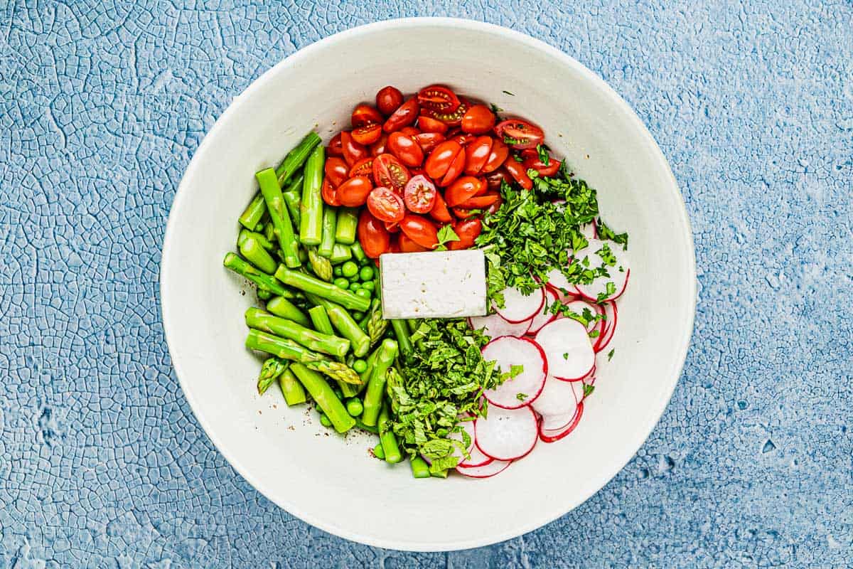 Herbs, sliced radishes, halved cherry tomatoes, peas, asparagus, and feta cheese in a large bowl before being mixed.