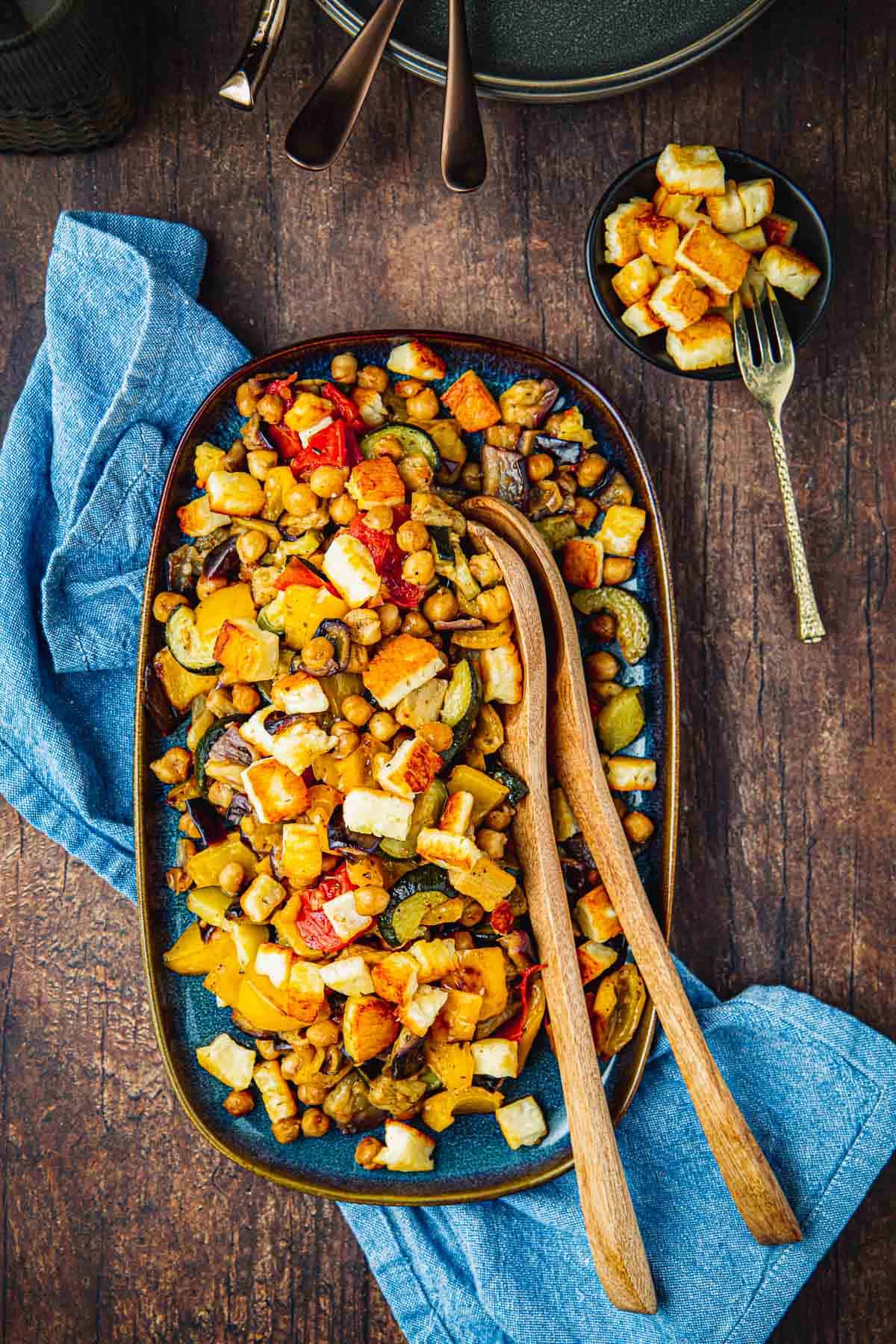 Overhead shot of Roasted Vegetable Salad on a blue serving dish with two wooden serving spoons, a blue napkin, and more fried cheese on the side.