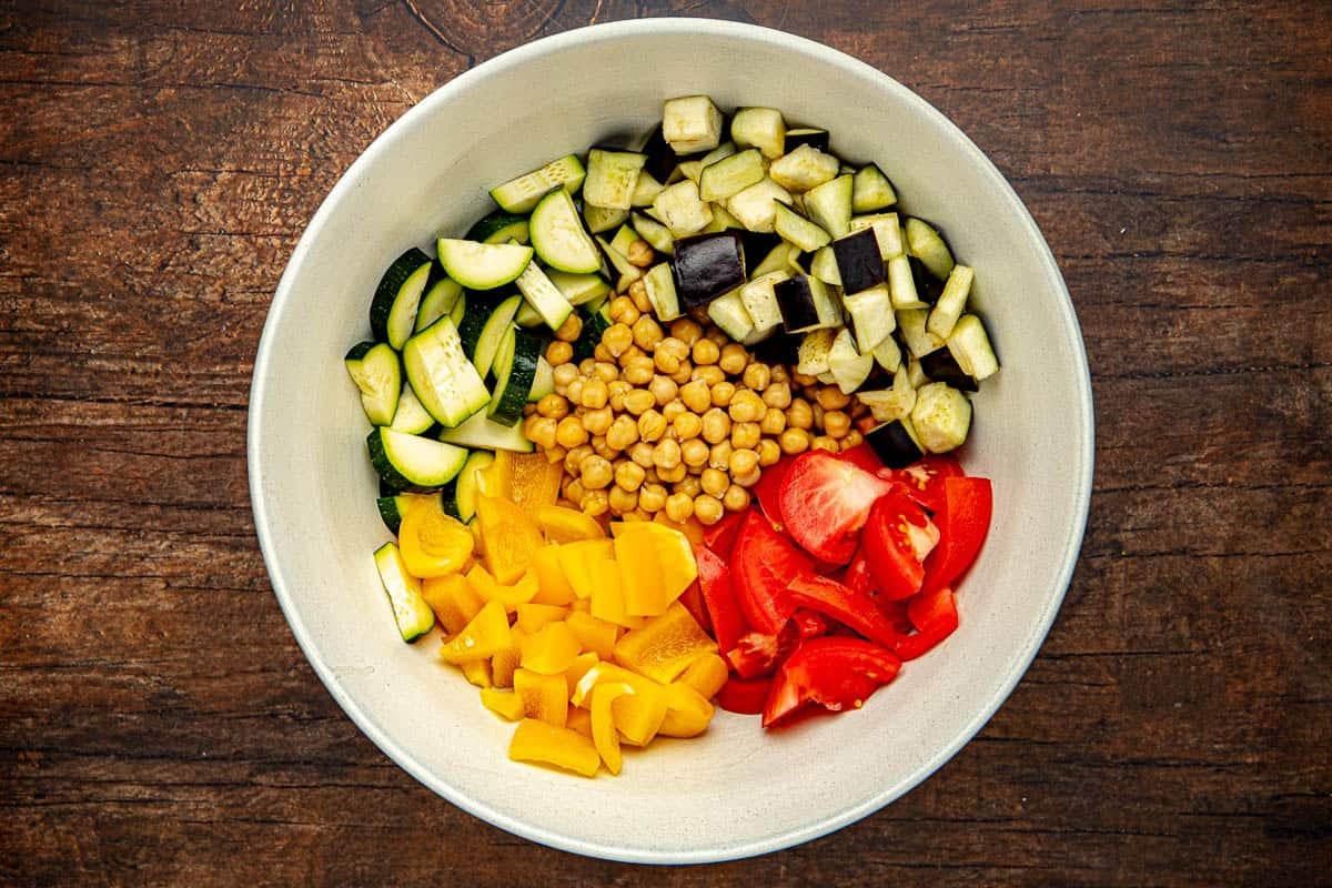 Raw chickpeas, zucchini, eggplant, tomatoes, and yellow bell peppers in a bowl before being mixed.