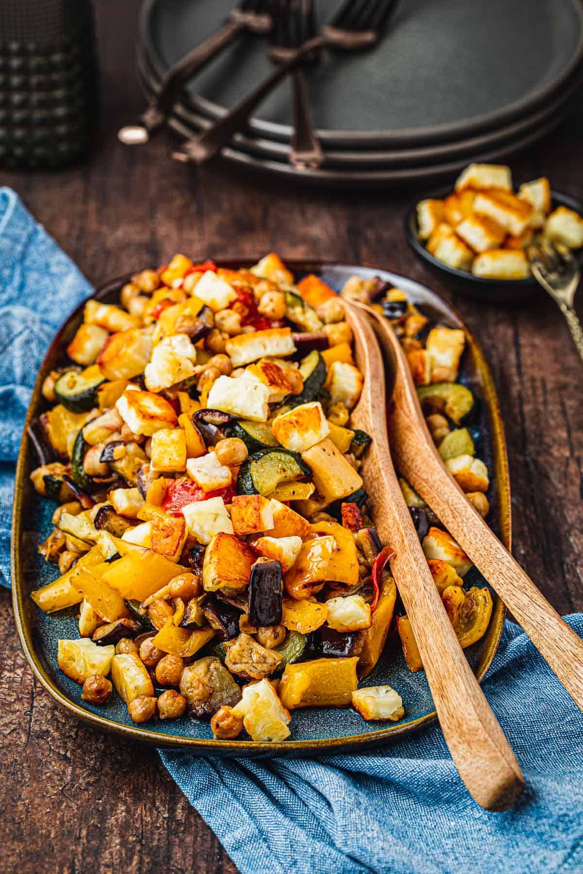 Side shot of Roasted Vegetable Salad with fried Halloumi on top, two wooden serving spoons, and a blue napkin.