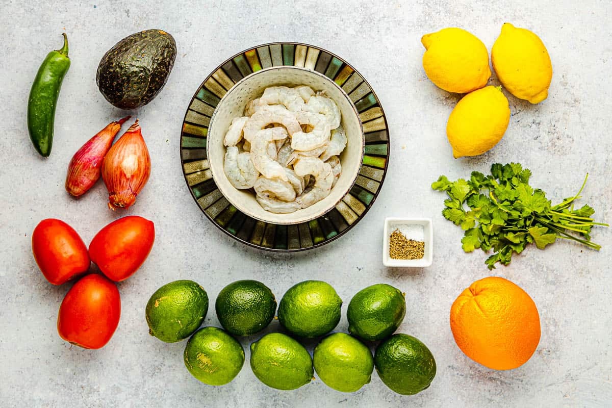 ingredients of shrimp ceviche including shrimp, lemons, limes, an orange, avocado, cilantro, tomatoes, shallots a jalapeno, salt and pepper.