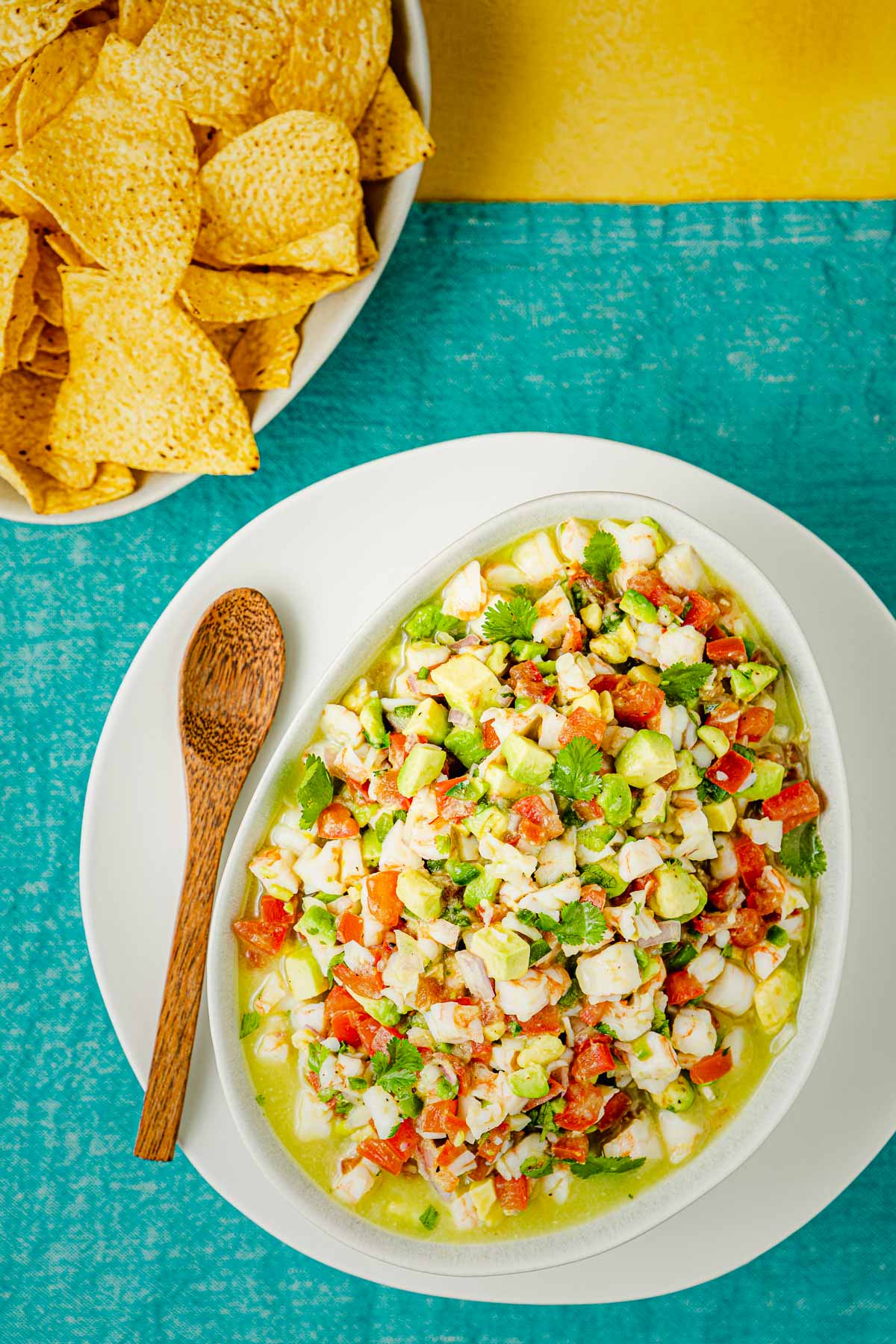 a bowl of shrimp ceviche on a plate next to a wooden spoon with a bowl of tortilla chips in the background.