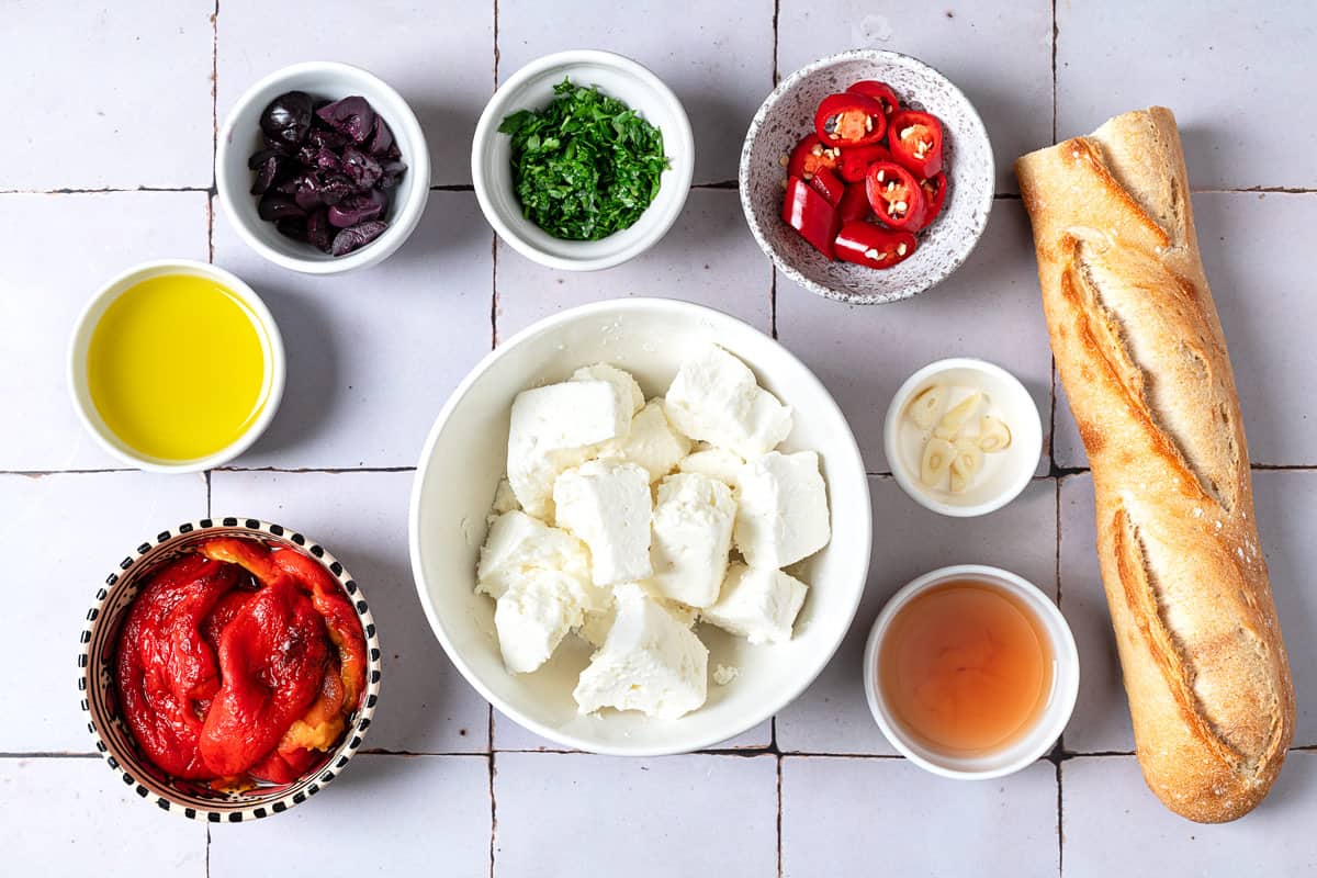 Ingredients for Tirokafteri Spicy Greek Feta Dip, including feta cheese, roasted red peppers, garlic, olive oil, parsley, olives, red wine vinegar, and red chili pepper.