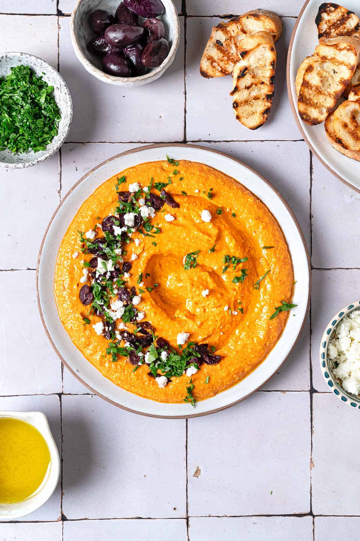 Tirokafteri Spicy Greek Feta Dip with grilled bread on the side and a feta, parsley, and olive garnish in a half moon shape.