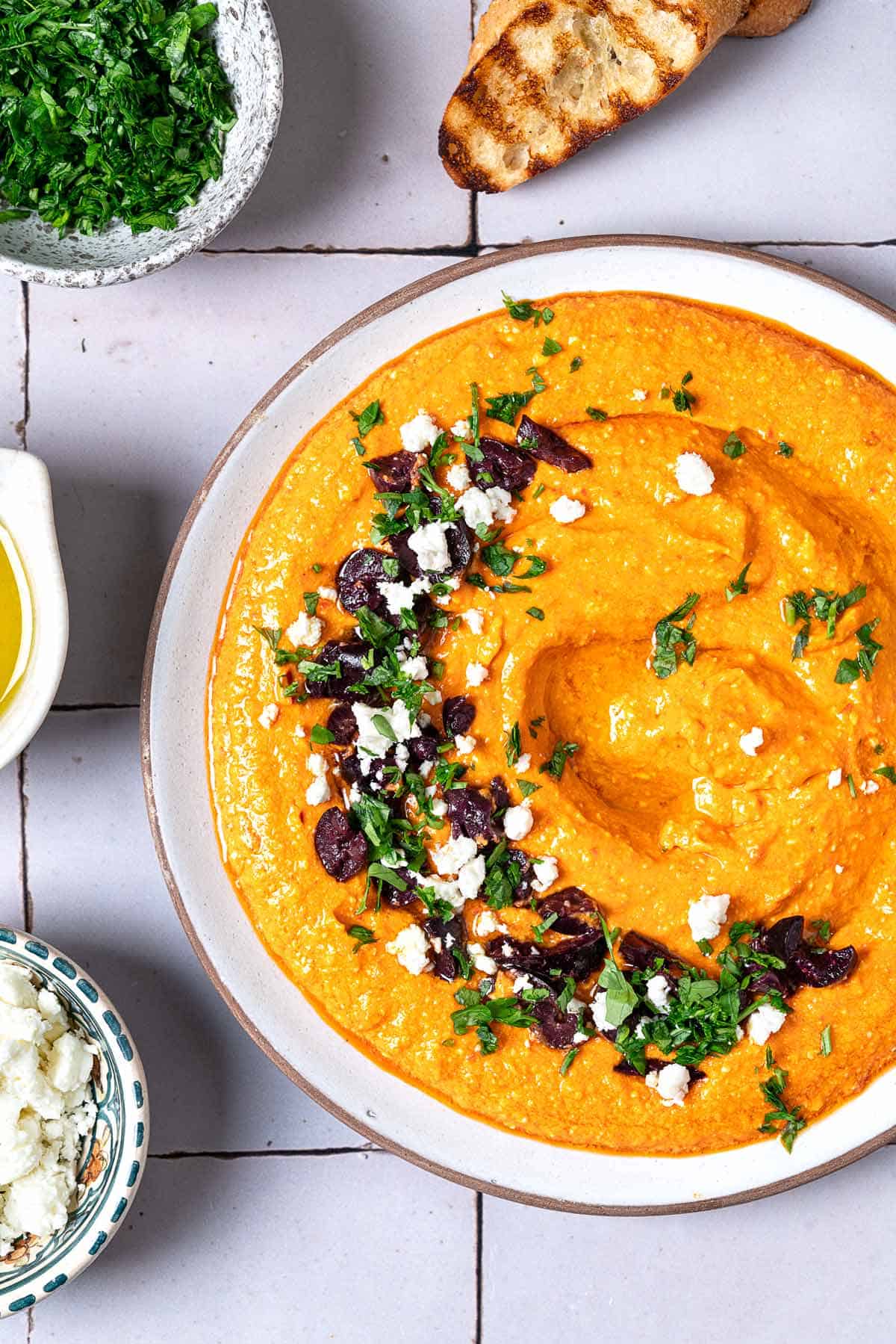 Close up shot of Tirokafteri Spicy Greek Feta Dip with grilled bread on the side and a feta, parsley, and olive garnish in a half moon shape.