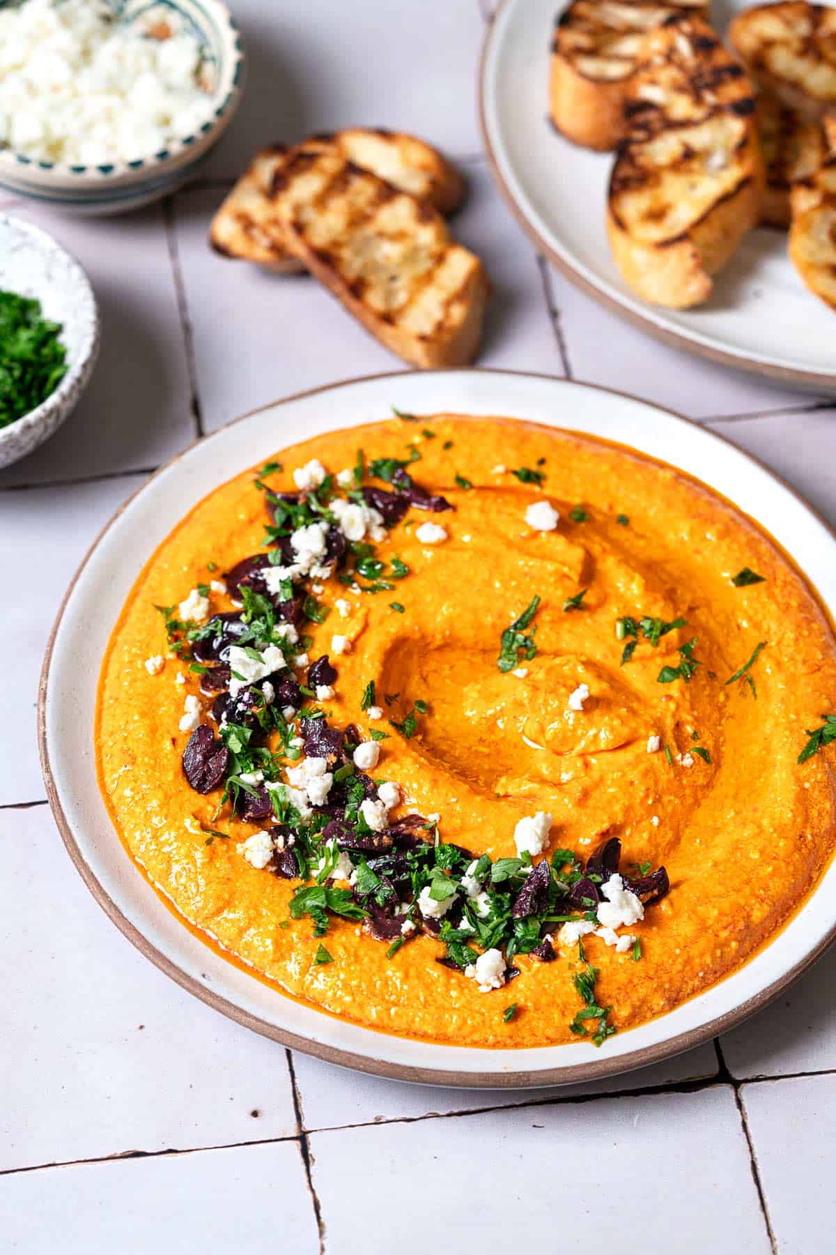 Side shot of Tirokafteri Spicy Greek Feta Dip with a feta, parsley, and olive garnish in a half moon shape and grilled bread in the background.