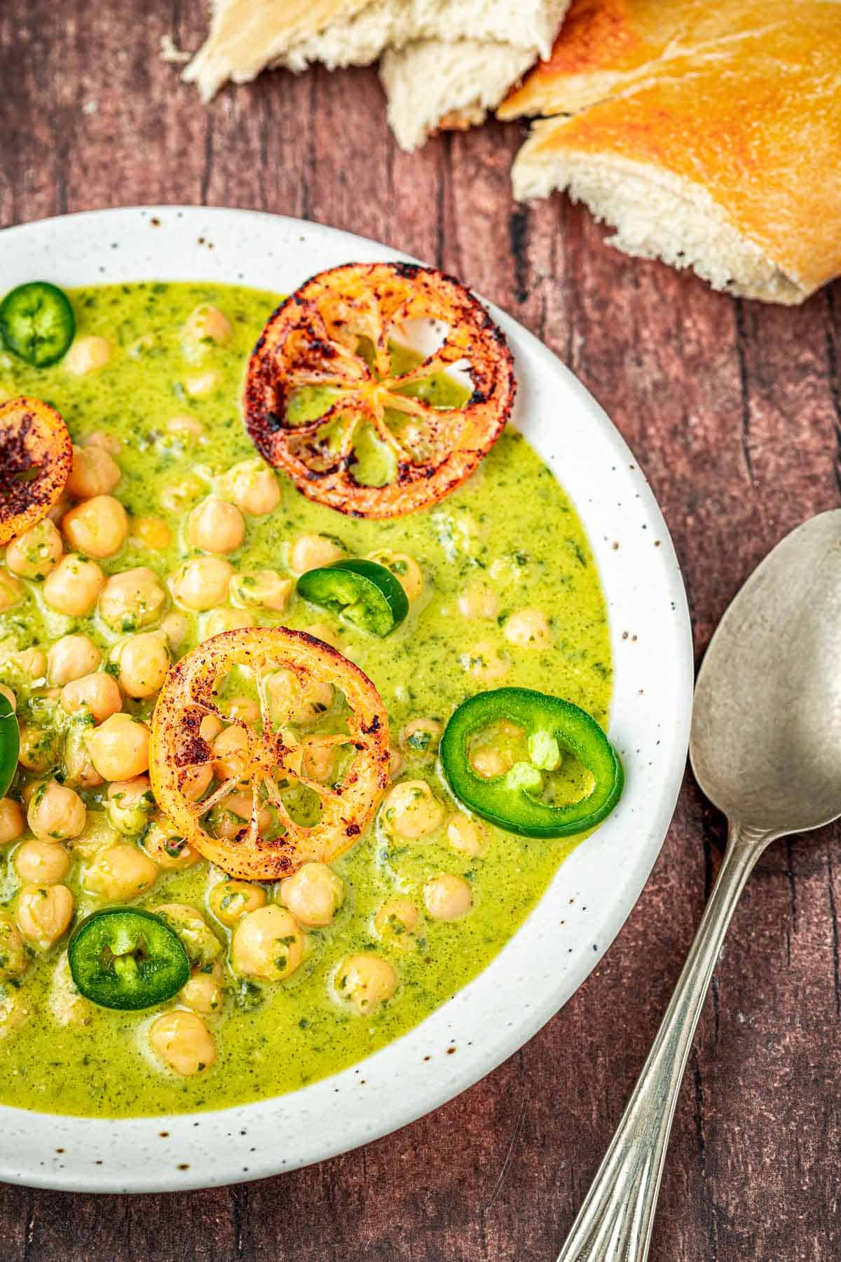 close up of a bowl of creamy pesto braised chickpeas topped with jalapeno and fried lemon slices next to a spoon and pieces of crusty bread.