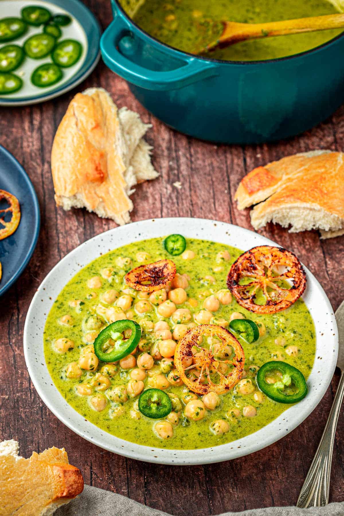a bowl of creamy pesto braised chickpeas topped with sliced jalapenos and fried lemons next to a spoon, pieces of crusty bread a pot of braised chickpeas and a plate of sliced jalapenos.