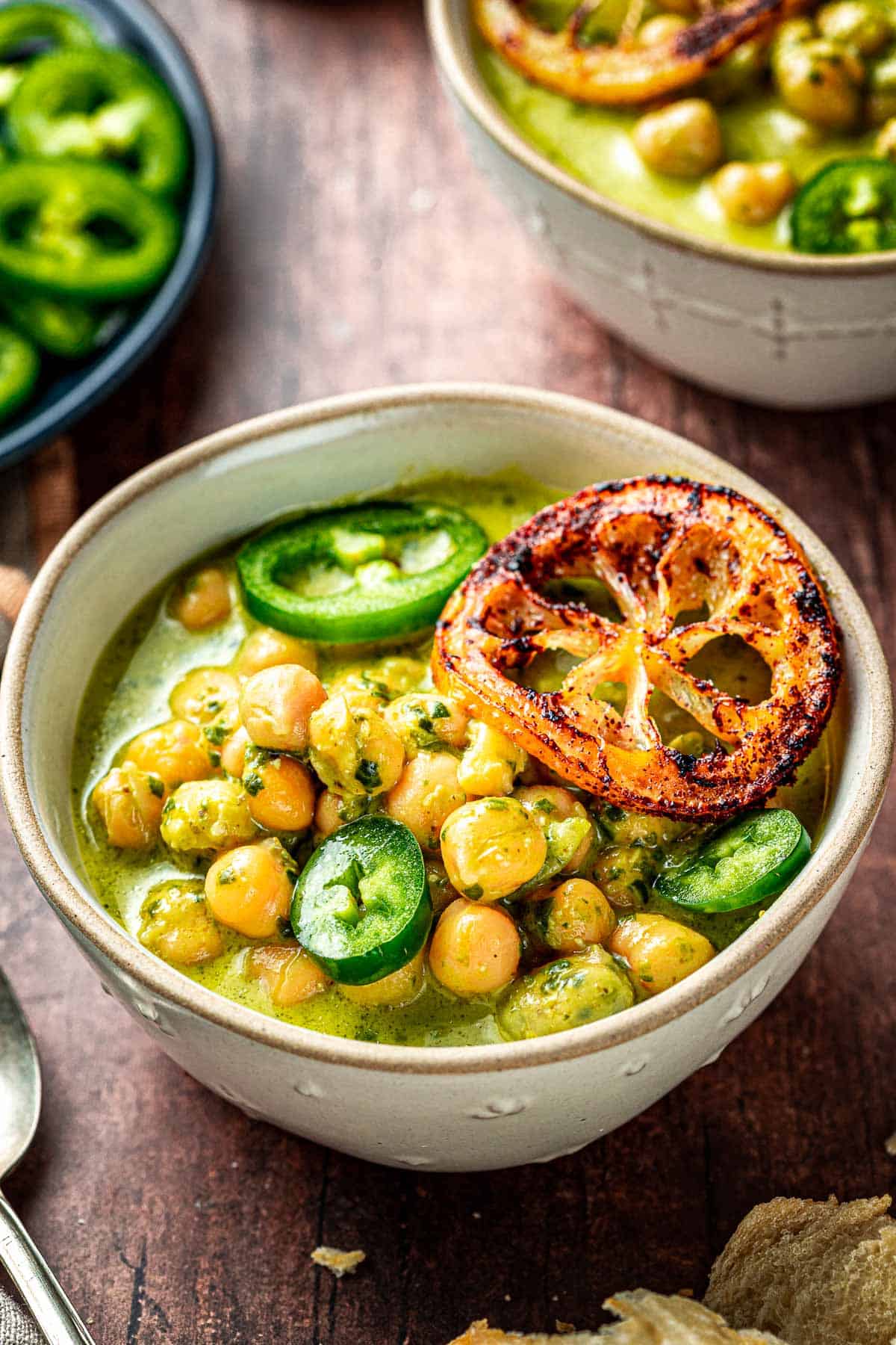 gros plan de deux bols de pois chiches braisés au pesto crémeux garnis de jalapeno et de tranches de citron frites, à côté d'une assiette de tranches de jalapeno.