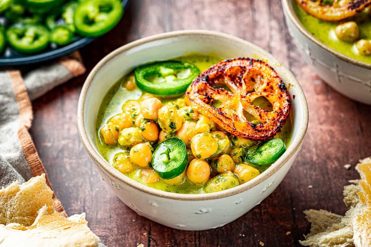 close up of two bowls of creamy pesto braised chickpeas topped with jalapeno and fried lemon slices next to a plate of sliced jalapenos.