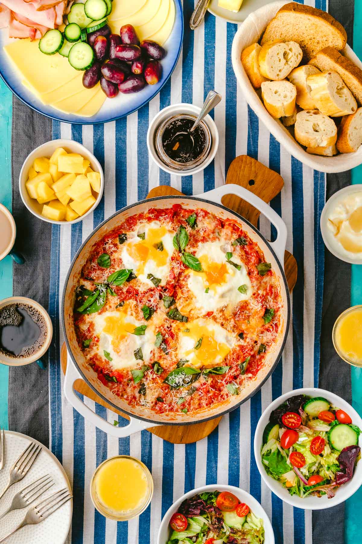 a skillet of eggs in purgatory next to a meat and cheese tray, a basket of sliced crusty bread, jam in a jar, a bowl of pineapple, a cup of coffee, a cup of orange juice, 2 bowls of salad, a bowl of yogurt, and a plate of forks.