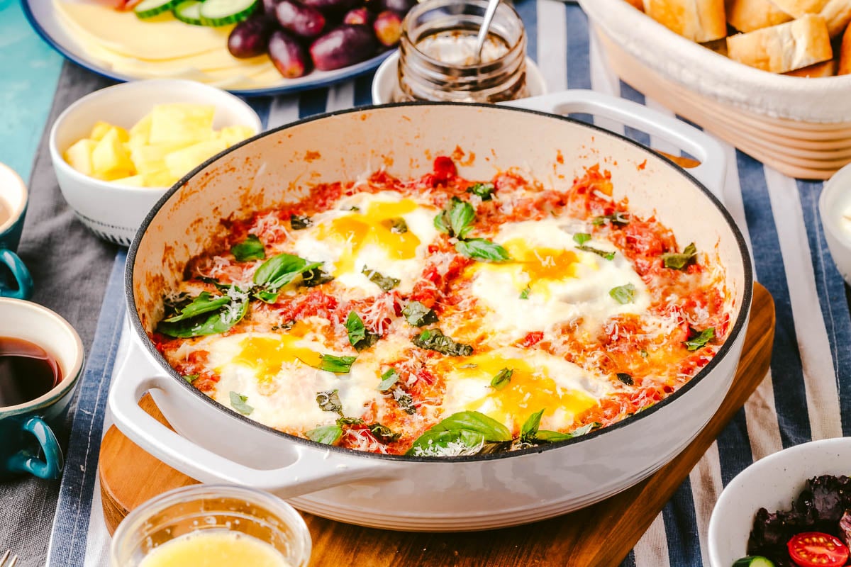 a skillet of eggs in purgatory next to a meat and cheese tray, a basket of sliced crusty bread, jam in a jar, a bowl of pineapple, a cup of orange juice, and a bowl of salad.