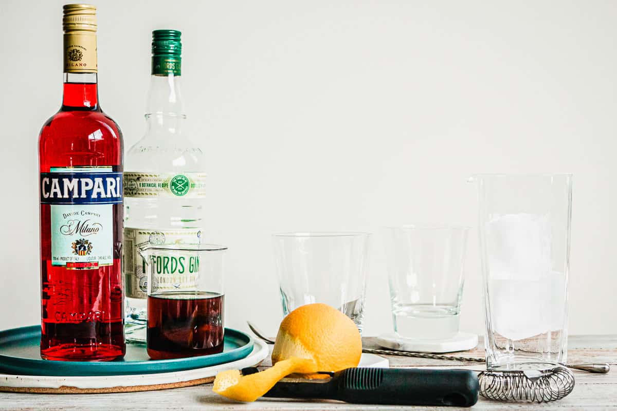 A negroni cocktail in a glass cocktail mixer with a bottle of campari, gin, and a peeled orange on the side.