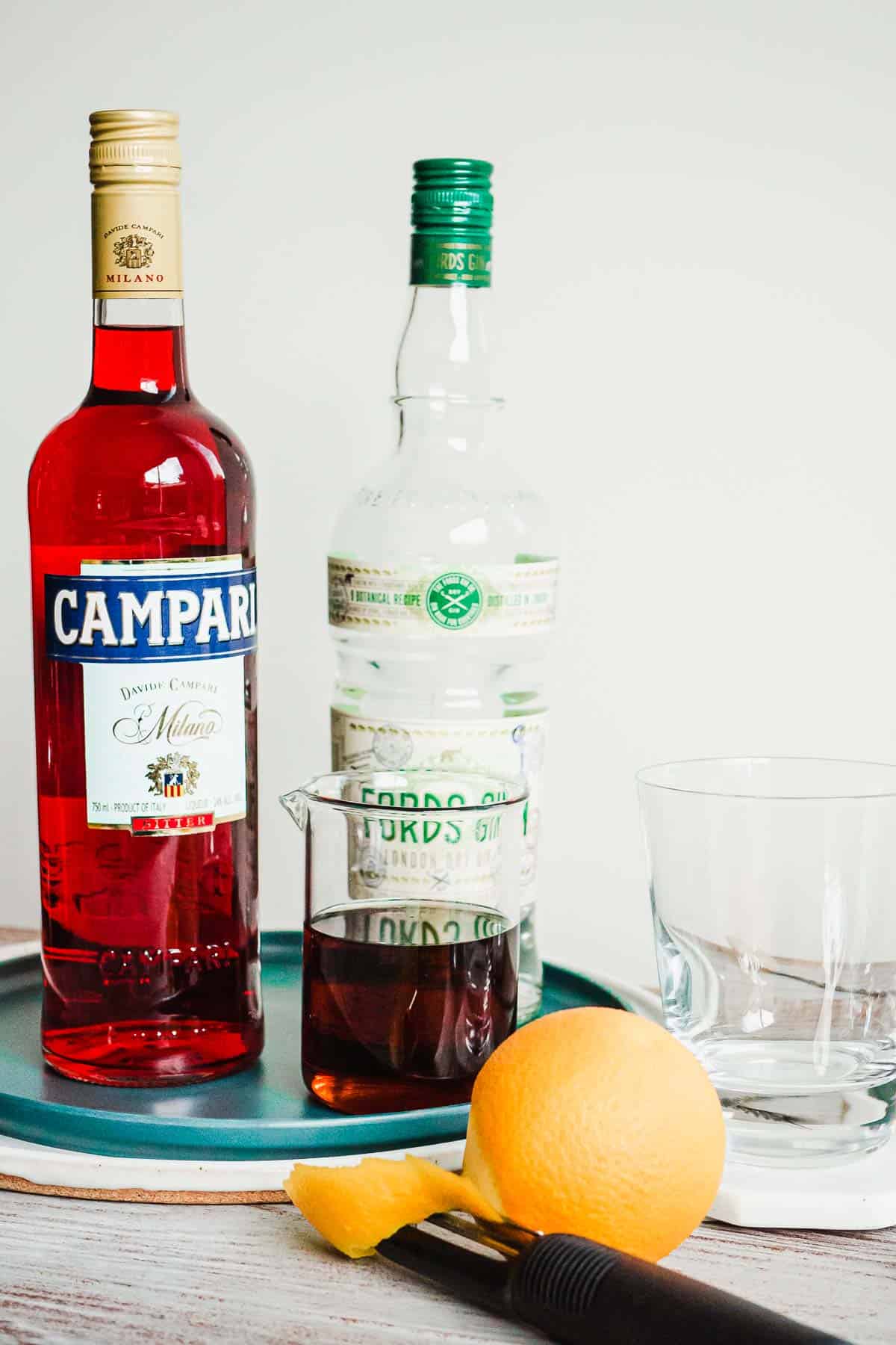 A negroni cocktail in a glass cocktail mixer with no ice and a bottle of campari, gin, and a peeled orange on the side.