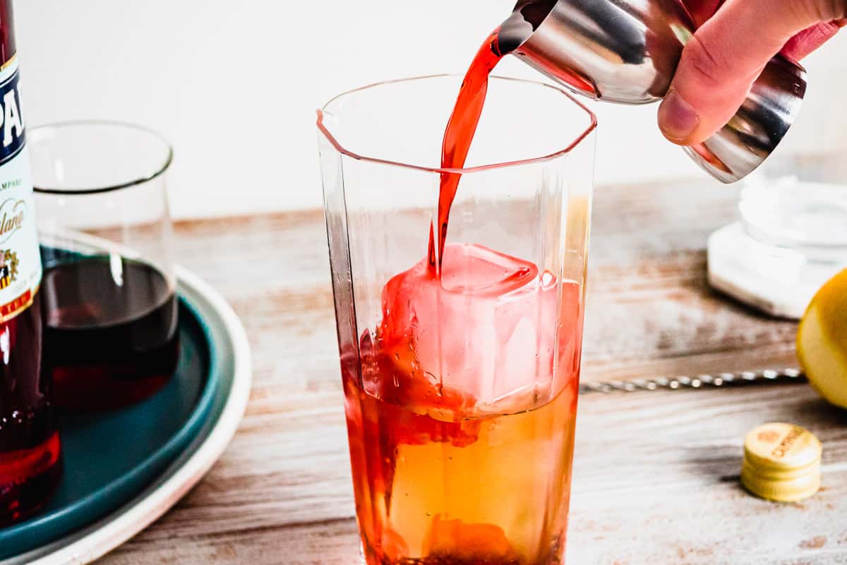 A hand pours campari into a glass filled with ice.