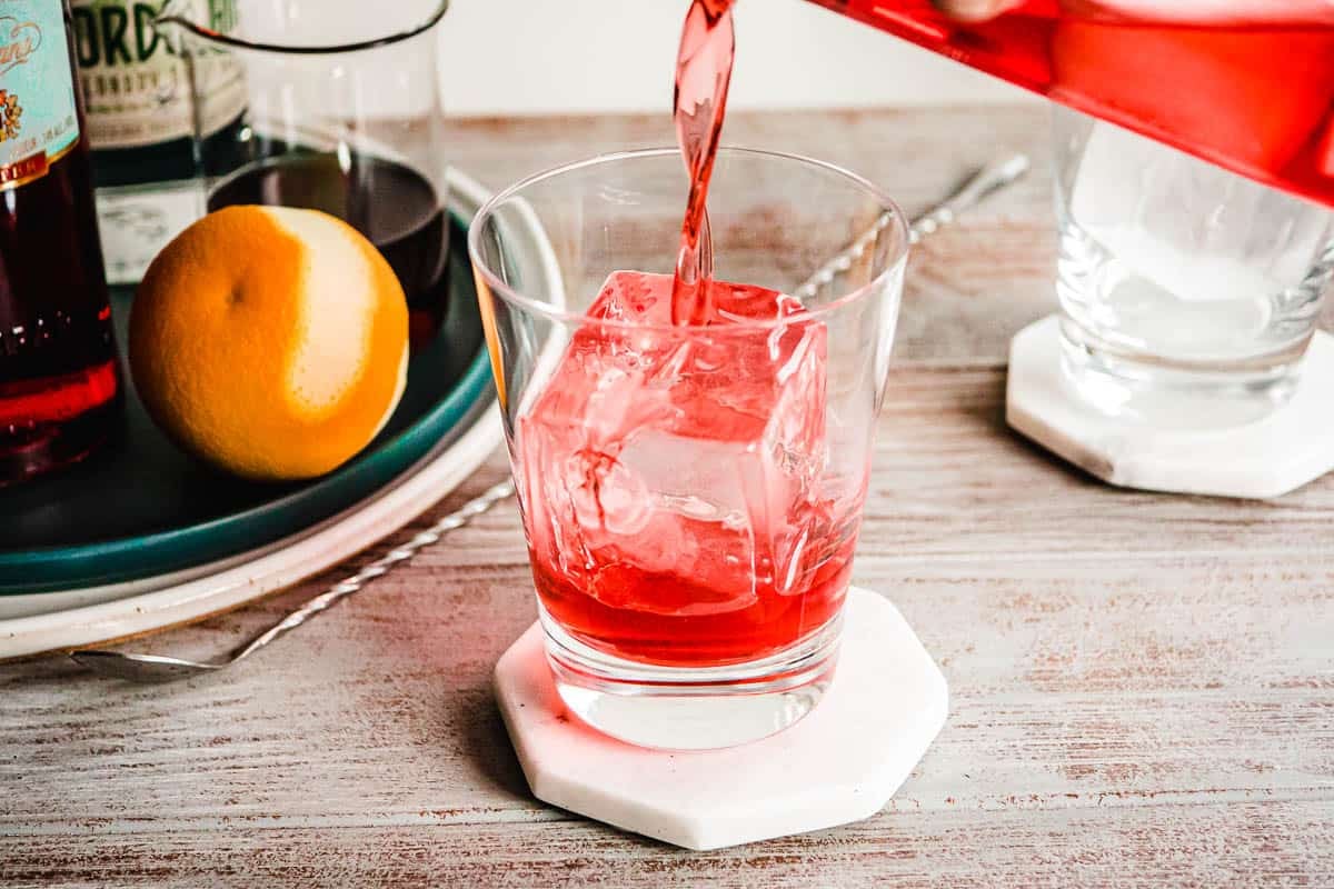 A person pours a negroni into a glass with one large ice cube.