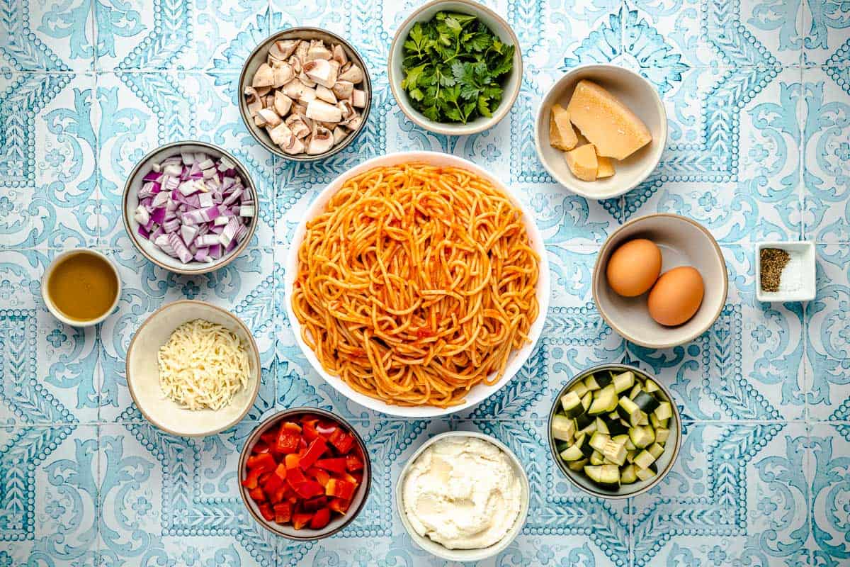 ingredients for spaghetti pie including leftover spaghetti, parsley, mushrooms, red onion, olive oil, mozzarella cheese, red pepper, ricotta, zucchini, eggs, salt, pepper and parmesan cheese.