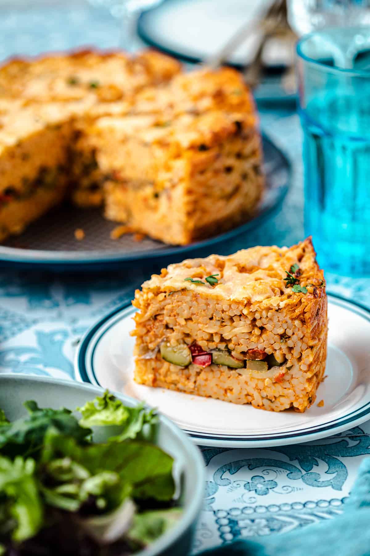 A slice of spaghetti pie on a plate next to an entire spaghetti pie, glass of water and a bowl of salad.