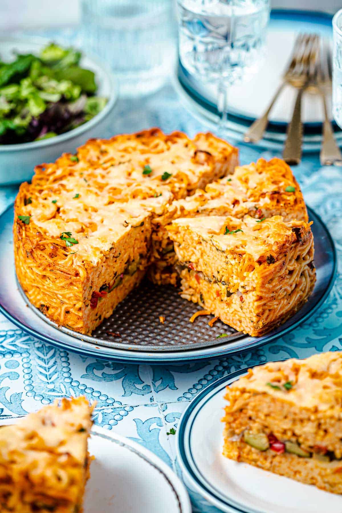 spaghetti pie on a plate next to a glass of water, a bowl of salad and two slices of spaghetti pie on plates.