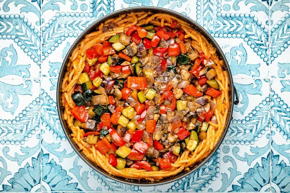 spaghetti pie topped with roasted vegetables in a spring form pan before baking.