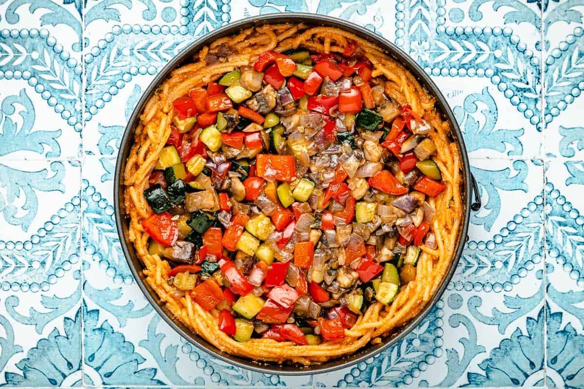 tarte aux spaghettis garnie de légumes rôtis dans un moule à charnière avant la cuisson.