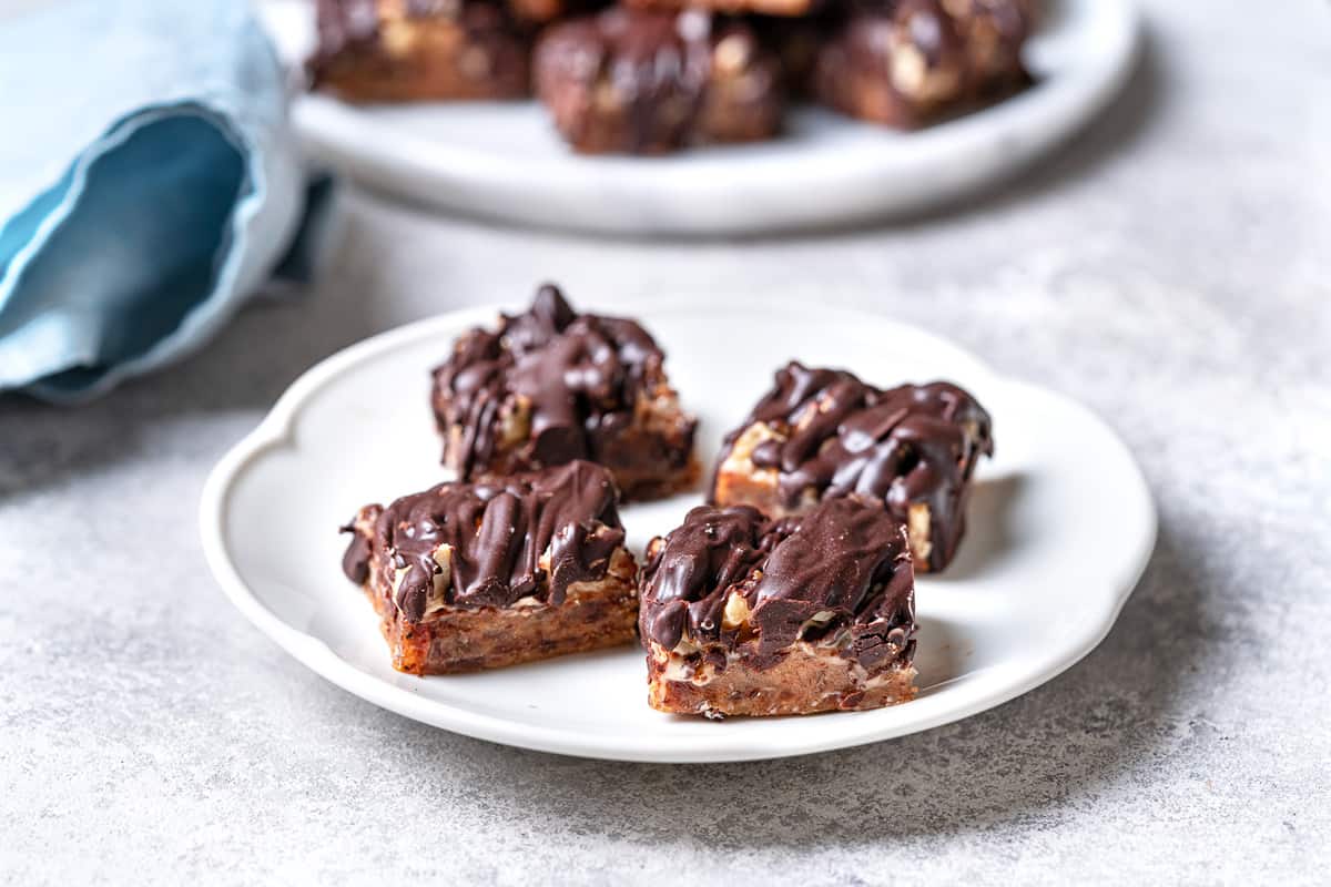 a close up of 4 chocolate date bars on a plate in front of a serving plate full of chocolate covered date bars.