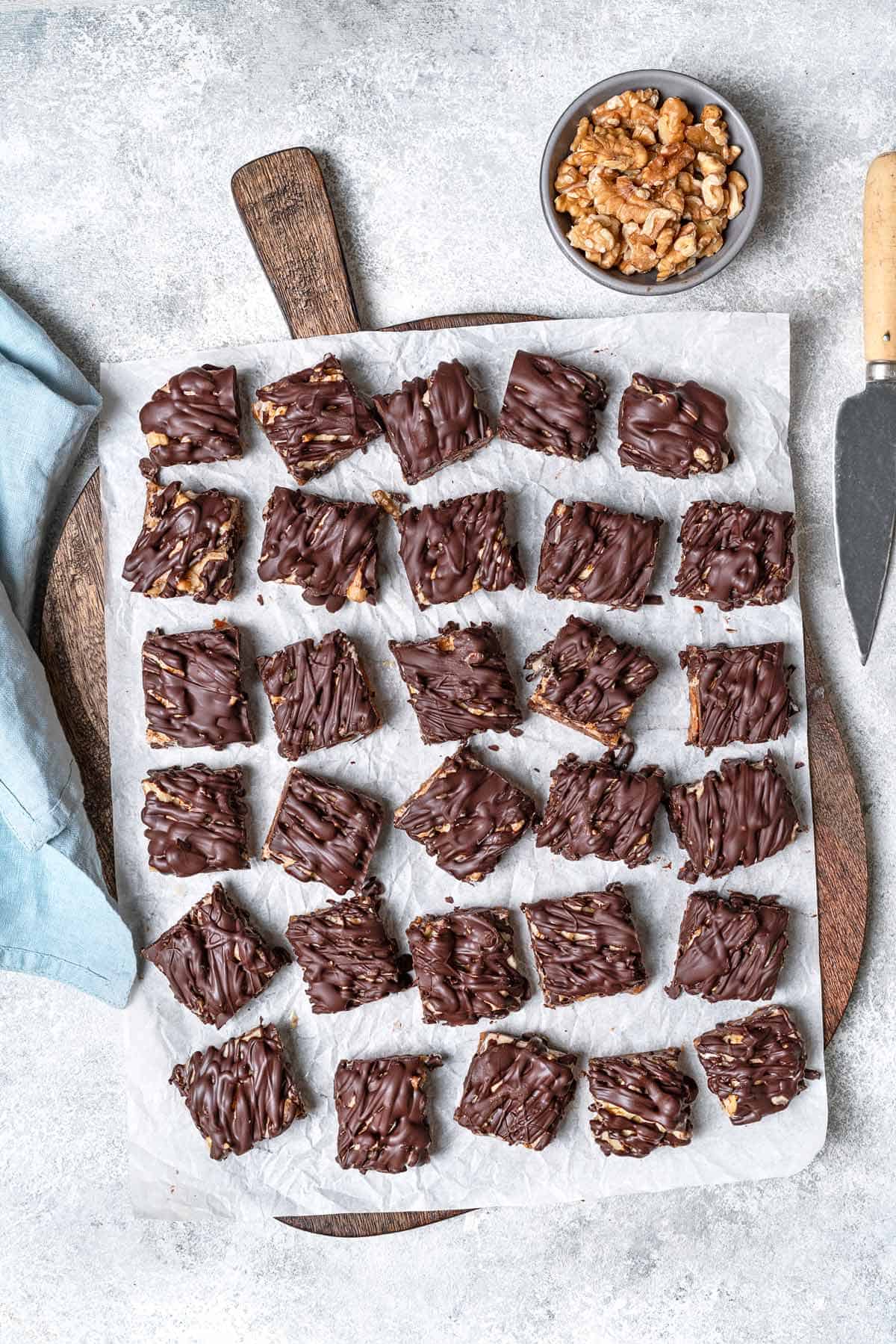 a whole batch of chocolate covered date bars on a parchment-lined serving tray next to a bowl of walnuts and a knife.