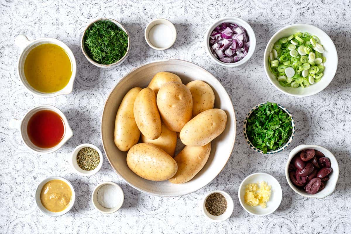 Ingredients for Greek Potato Salad, including yellow potatoes, salt, red onions, green onions, parsley, dill, Kalamata olives, red wine vinegar, Dijon mustard, dried oregano, garlic, salt, pepper, and olive oil.