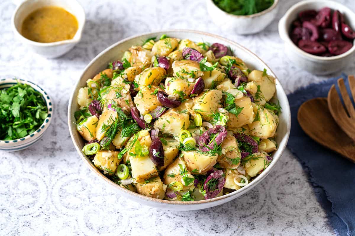 Side shot of a large serving bowl with greek potato salad and wooden serving spoons on the side.