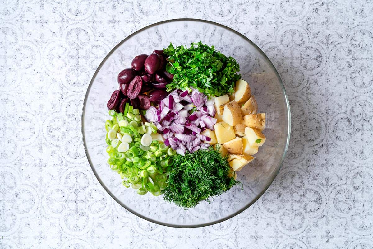 A bowl with sliced potatoes, onions, herbs, green onions, and olives.