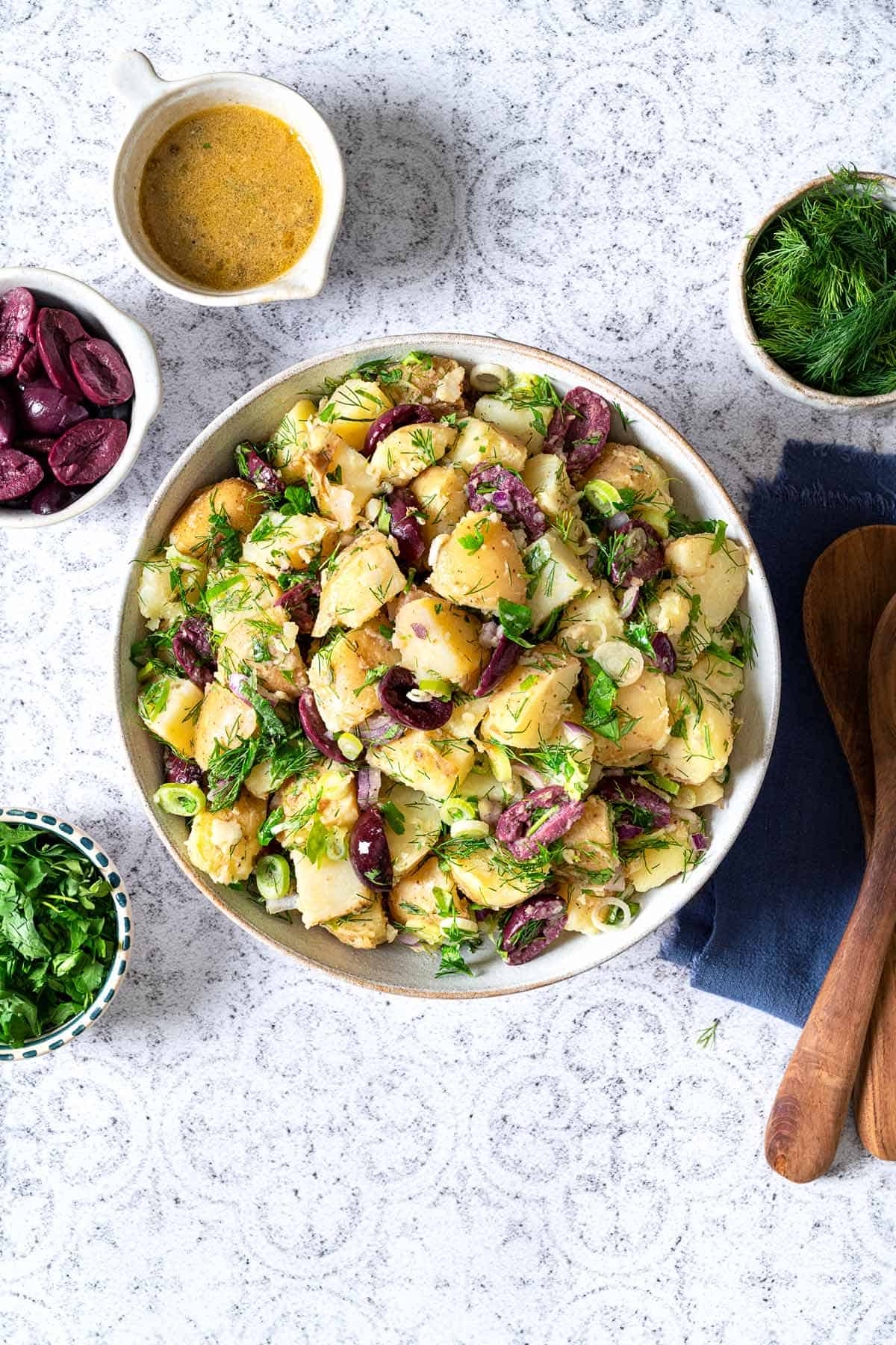 Bowl of greek potato salad with bowls of dressing, herbs, olives and two wooden spoons on the side.