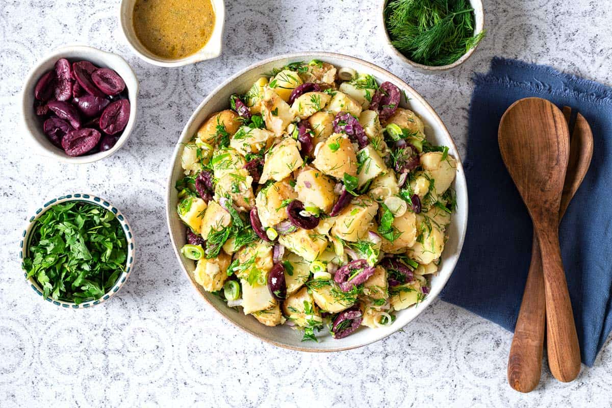 Overhead shot of a large bowl of greek potato salad with bowls of dressing, herbs, olives and two wooden spoons on the side.