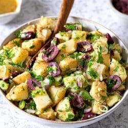 Side shot of a large bowl of greek potato salad with a wooden serving spoon.