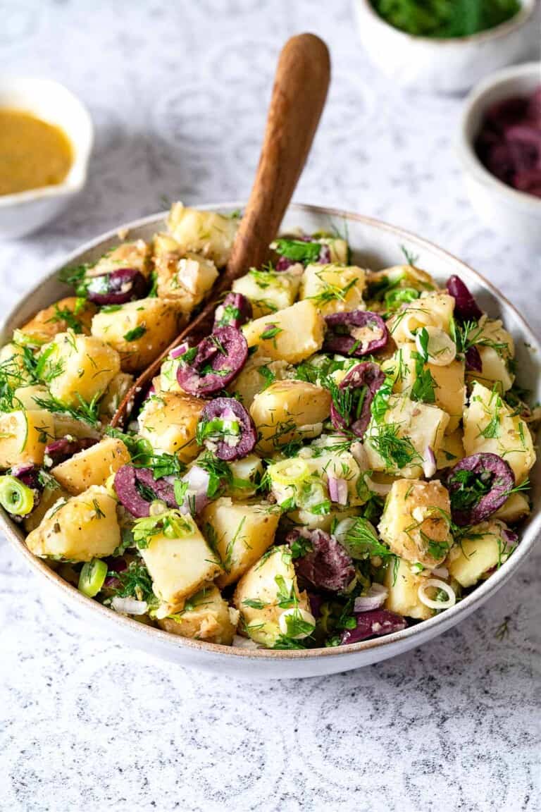 Side shot of a large bowl of greek potato salad with a wooden serving spoon.