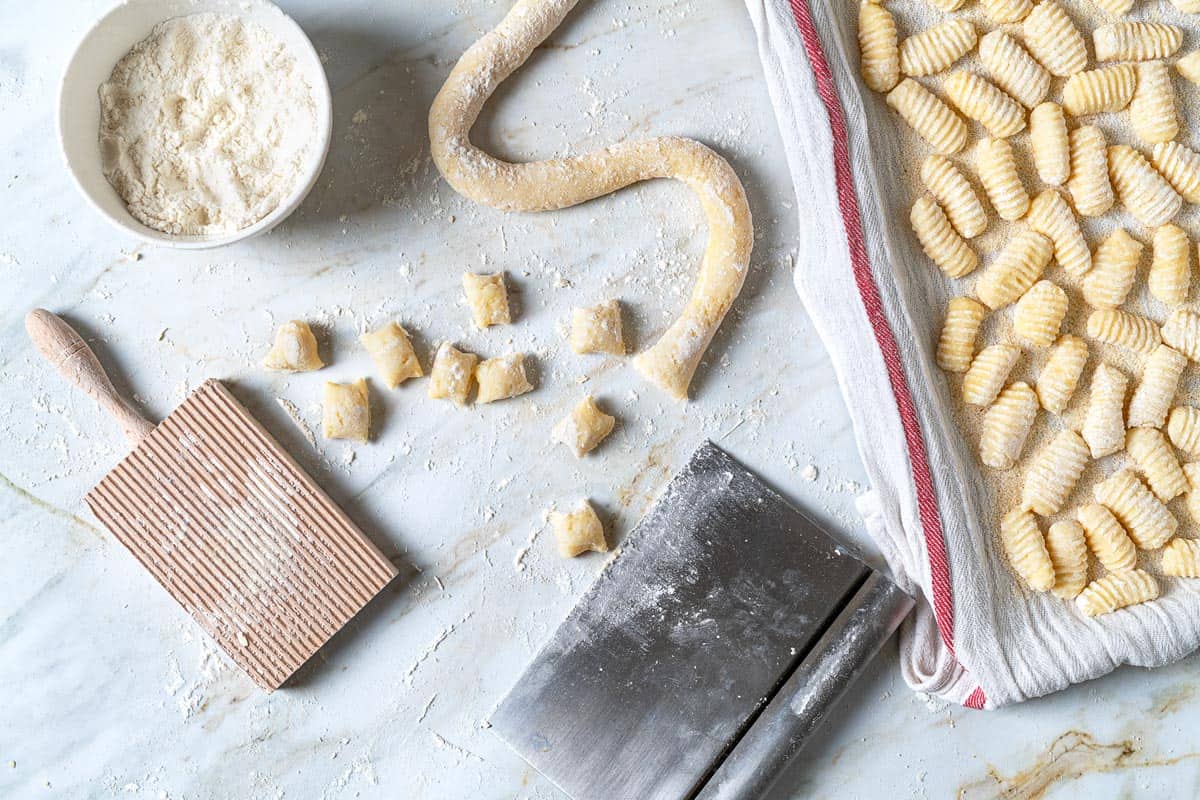 A rope of potato gnocchi that has been cut into small dumplings, with a tray of shaped potato gnocchi, a gnocchi board, bowl of semolina flour, and a bench scraper on the side.