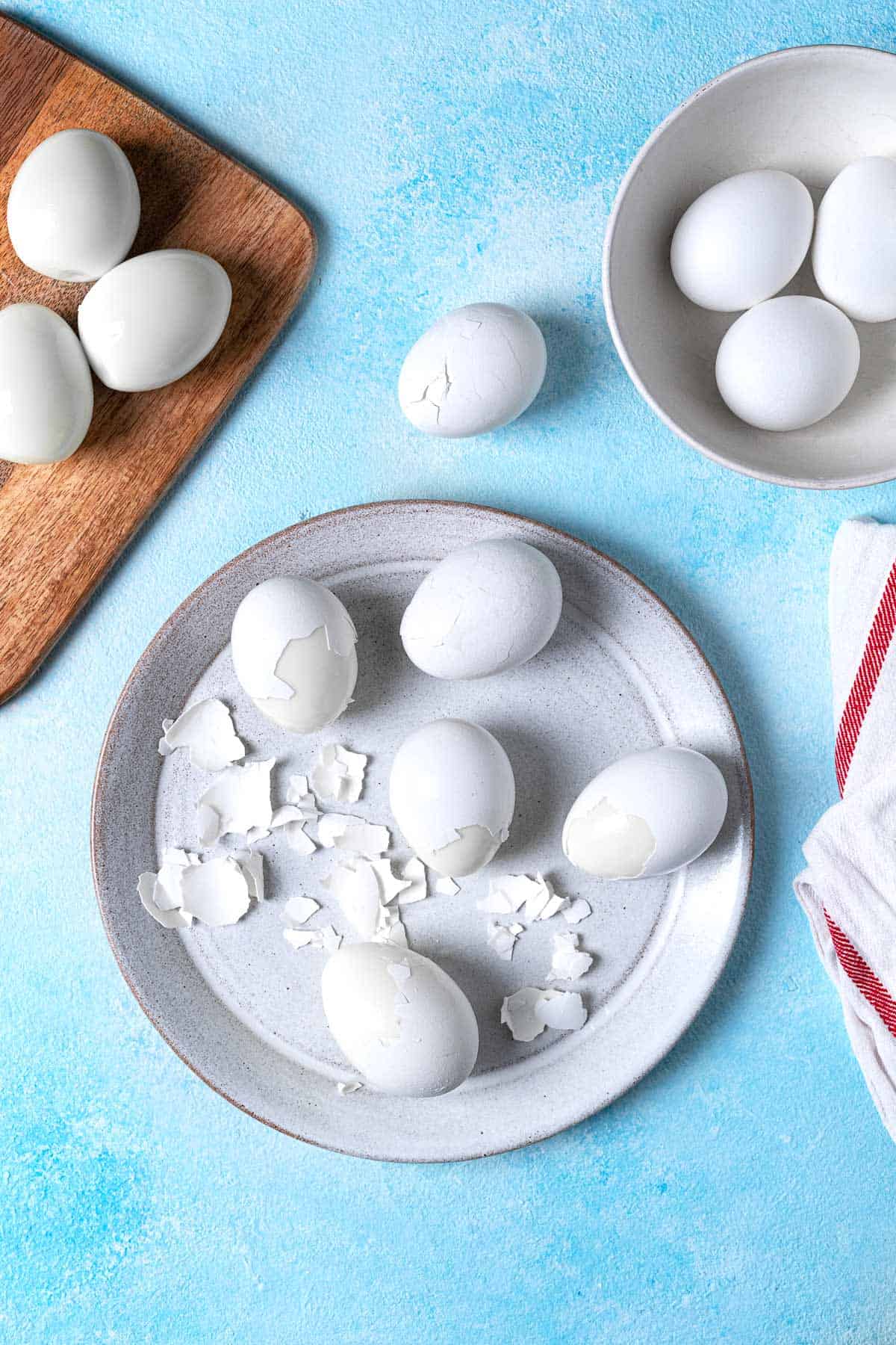 5 partially peeled hard boiled eggs on a plate next to a wooden tray with3 fully peeled eggs and a bowl with 3 unpeeled eggs.