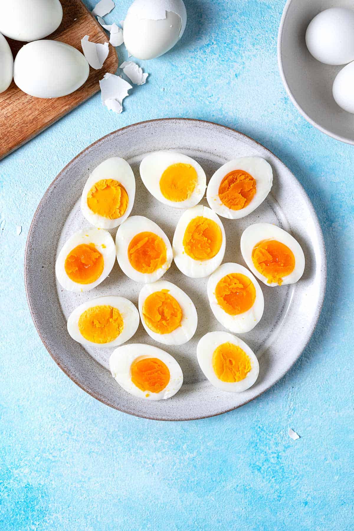 12 hard boiled egg halves on a plate next to a bowl and a wooden tray with whole hard boiled eggs.