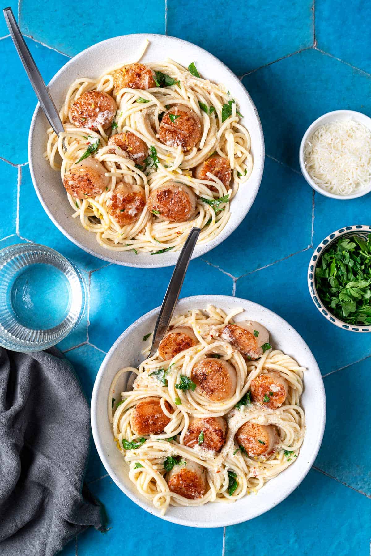 Two bowls of scallop pasta on a blue tile background with cheese and parsley on the side.