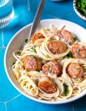 Bowl of pasta with scallops, lemon zest, and parsley on a blue tile background.