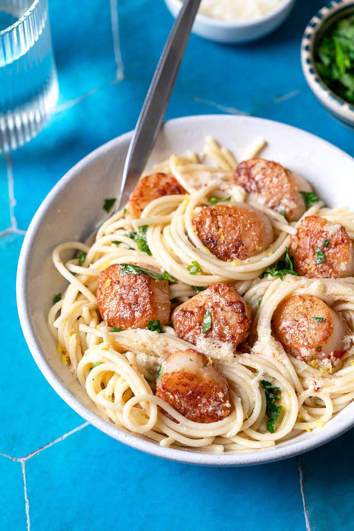 Bowl of pasta with scallops, lemon zest, and parsley on a blue tile background.