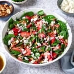 spinach strawberry salad topped with toasted almonds and feta in a bowl next to bowls of toasted almonds, mint and feta.