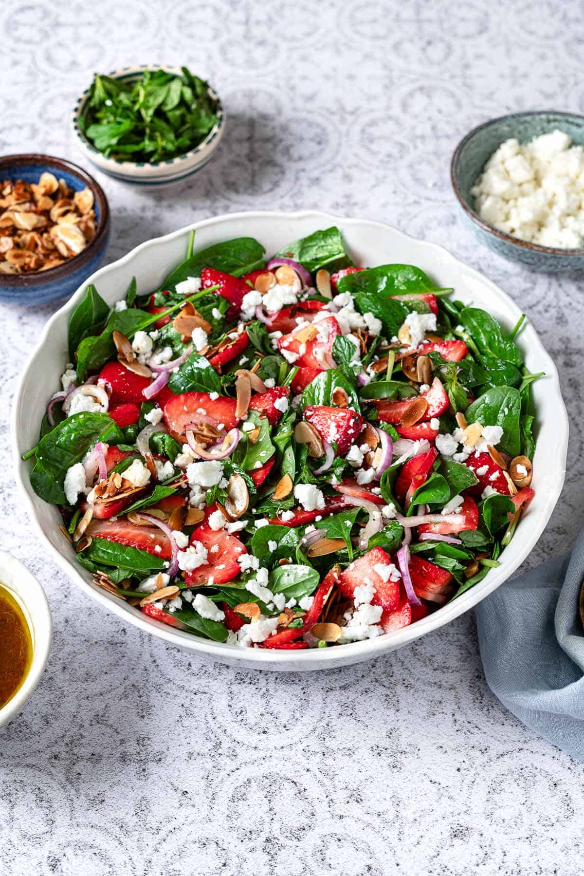 spinach strawberry salad topped with toasted almonds and feta in a bowl next to bowls of toasted almonds, mint and feta.