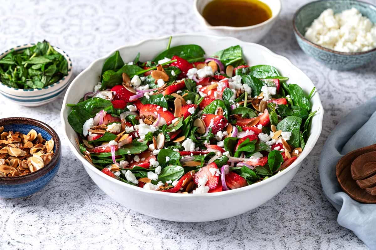 spinach strawberry salad topped with toasted almonds and feta in a bowl next to bowls of toasted almonds, mint, dressing and feta.