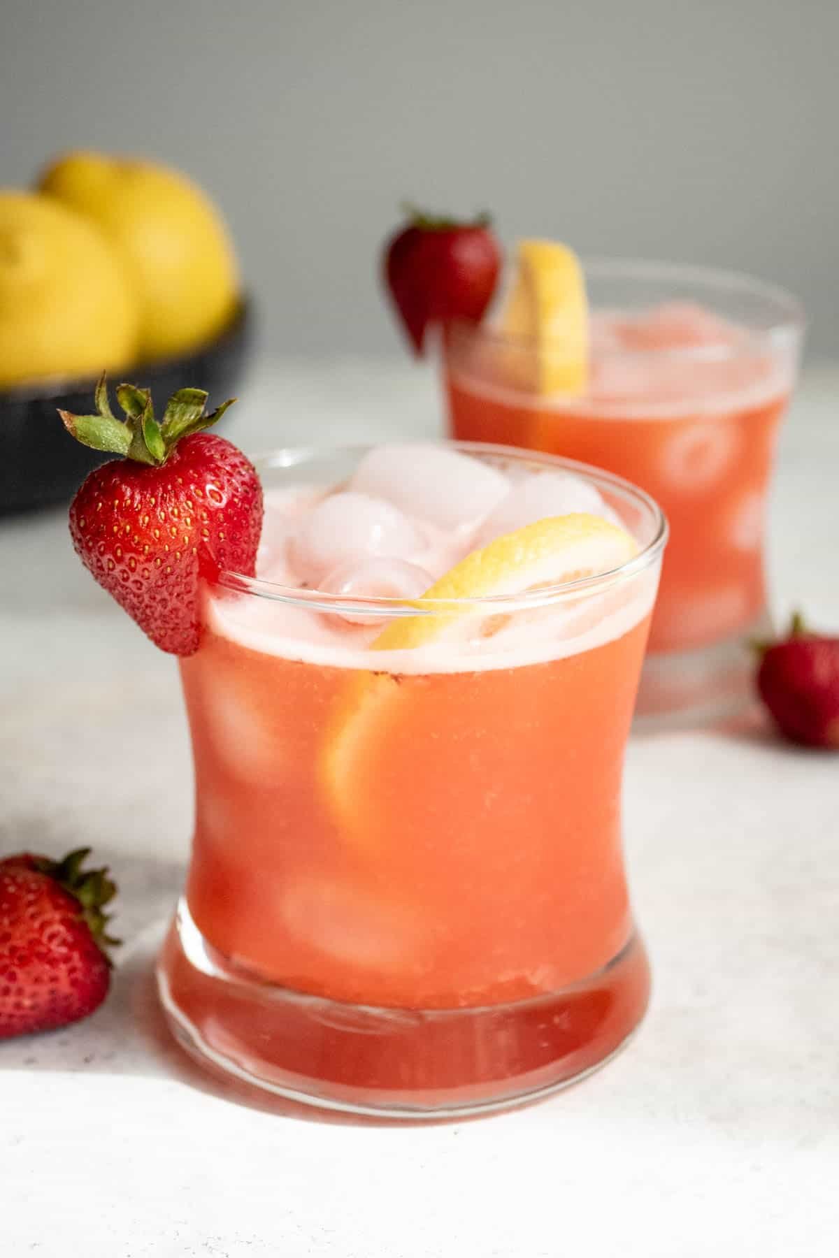 a glass of strawberry lemonade garnished with a strawberry and a lemon slice in front of another glass of strawberry lemonade and a bowl of lemons.