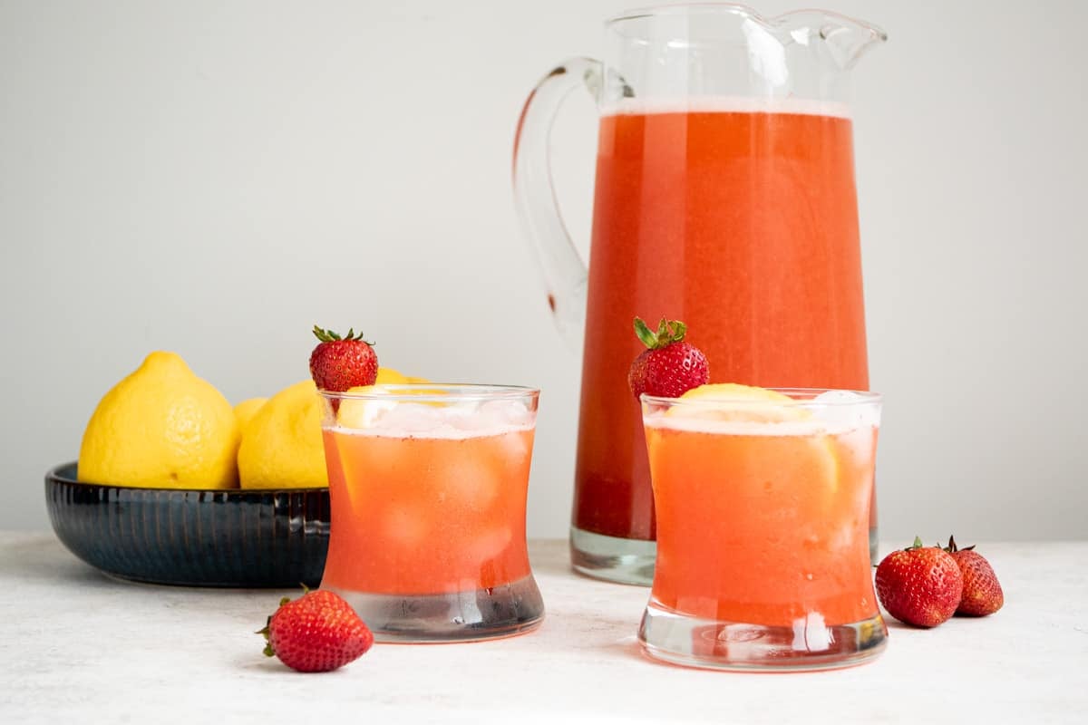 two glasses of strawberry lemonade garnished with strawberries and lemon slices in front of a pitcher of strawberry lemonade and a bowl of lemons.