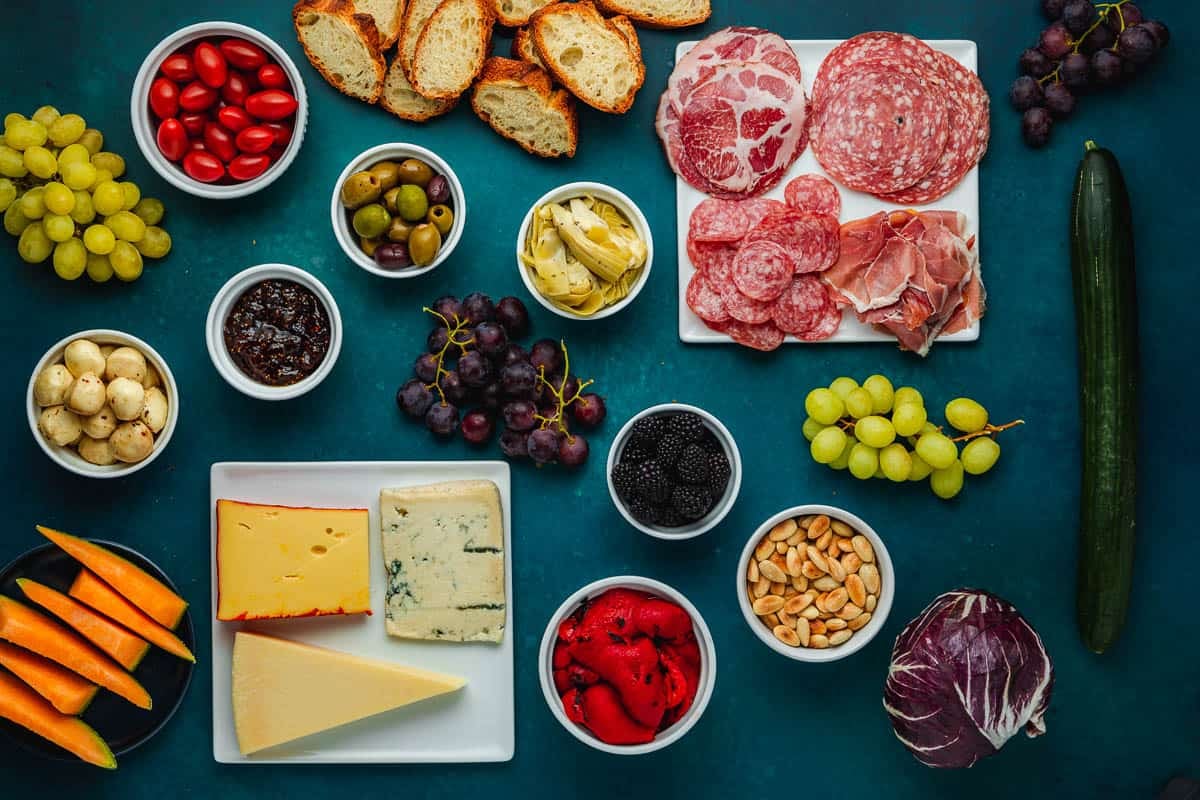 ingredients for an antipasto platter including slices of crusty bread, salami, prosciutto, capicola, grapes, cucumber, radicchio, blanched almonds, roasted red bell pepper slices, black berries, artichoke hearts, olives, fig jam, mozzarella balls, slices of cantaloupe, cherry tomatoes, gorgonzola cheese, fontina cheese and Pecorino Romano cheese.